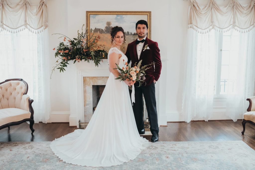 Bride in white dress, groom in red velvet suit jacket pose with flowers in front of fire place