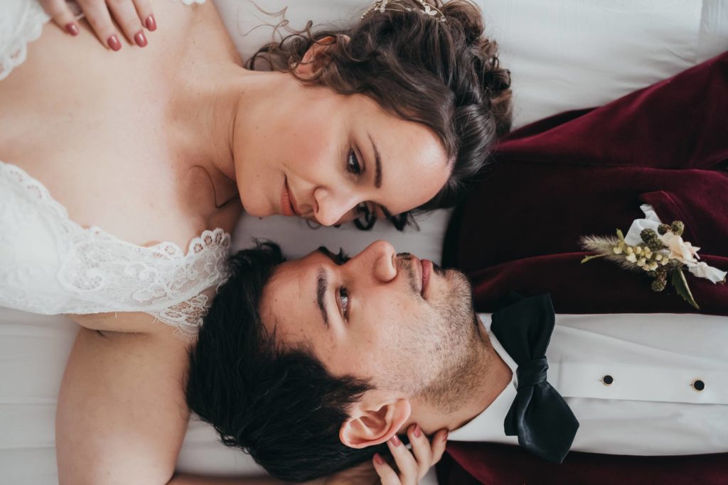 Bride and groom lay on bed with heads next to one another looking at each other