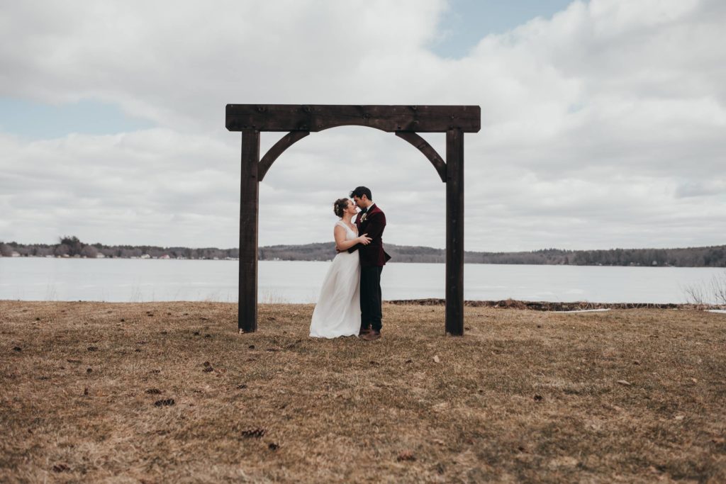 bride and groom standing under a wooden arch in front of a frozen lake